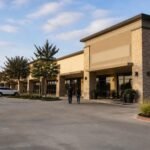 Illustration of a single-story suburban shopping center with tan stone pillars and dark storefront windows, a small café patio, young trees and a wide parking lot, as two people walk toward the entrance in early-morning light.