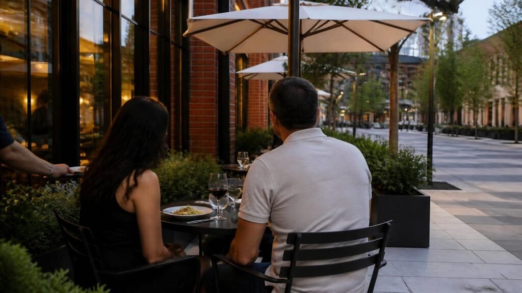 Illustration of a couple, seen from behind, sharing dinner and red wine at a bistro table on a brick restaurant's sidewalk patio under white umbrellas, with planters and a tree-lined paver street beyond.