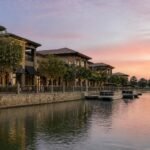 Illustration of a lakefront row of two-story beige stucco and stone-pillared buildings with dark awnings above a tan stone seawall and metal railing at sunset, with pontoon boats at a small dock and the pink sky reflected in calm water.
