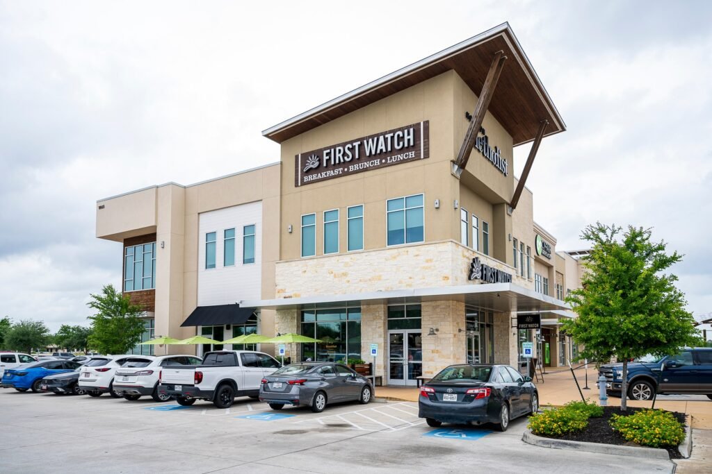 First Watch restaurant at The Boardwalk at Towne Lake in Cypress, a two-story tan stone and stucco building with green patio umbrellas out front