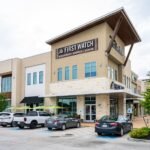 First Watch restaurant at The Boardwalk at Towne Lake in Cypress, a two-story tan stone and stucco building with green patio umbrellas out front