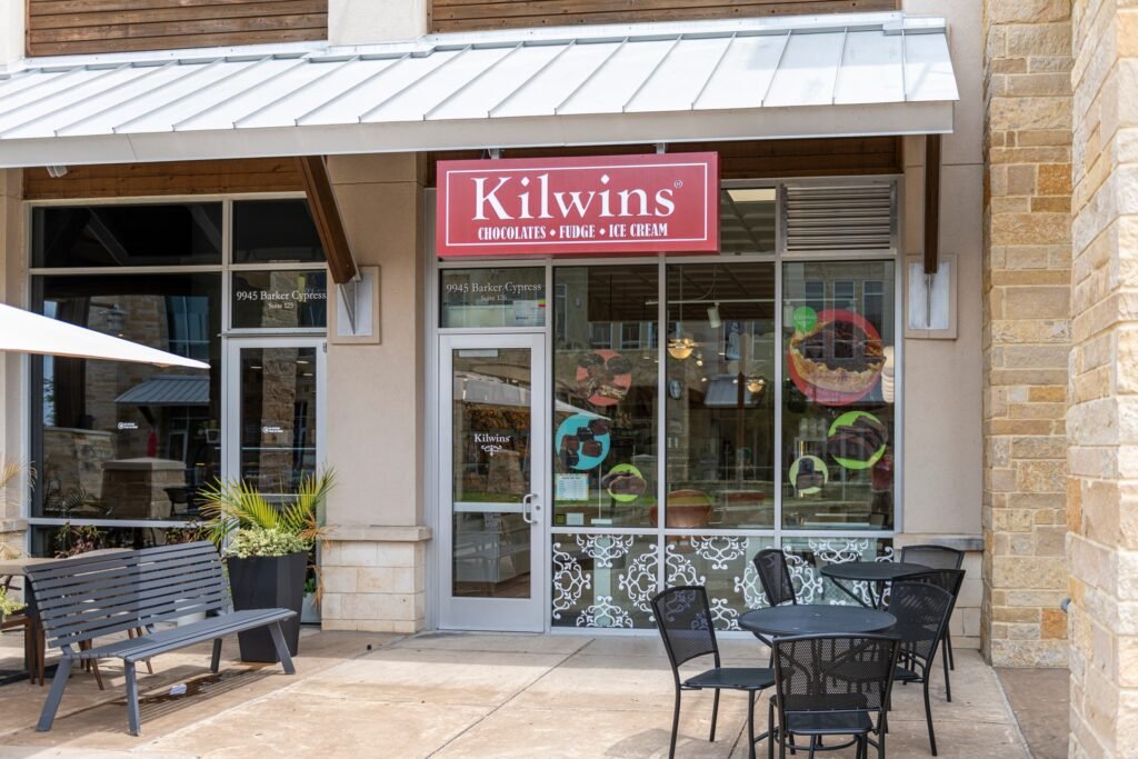 Kilwins chocolate, fudge and ice cream shop at The Boardwalk at Towne Lake, with a red sign, a metal awning and black café tables out front