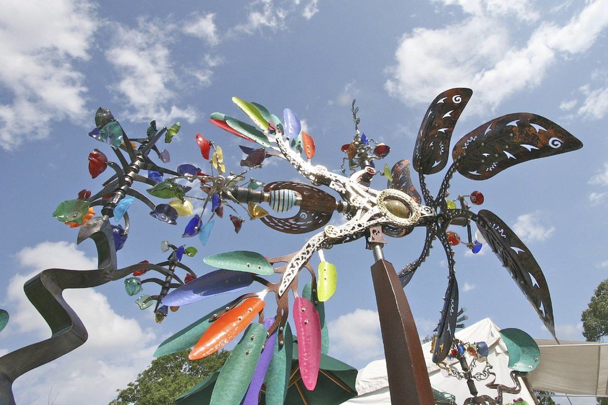 Colorful outdoor kinetic wind sculpture against sky with festival tent peak visible — licensed outdoor fine-art atmosphere from a prior Bayou City Arts Festival; not a documentary of named 2026 artists or Memorial Park 2026 booths