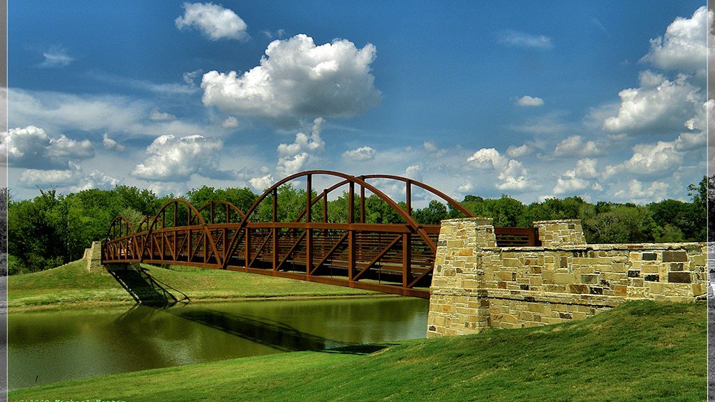Pedestrian footbridge spanning a lake at Bridgeland in Cypress, Texas, with grass banks, trees, and blue sky