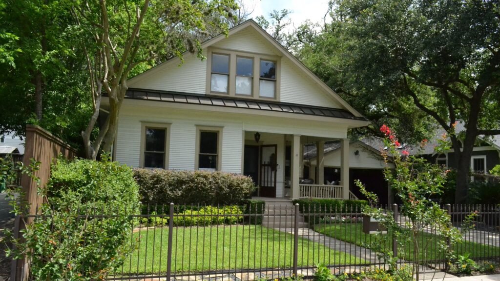 A white bungalow with a front porch behind an iron fence on a tree-lined Houston street