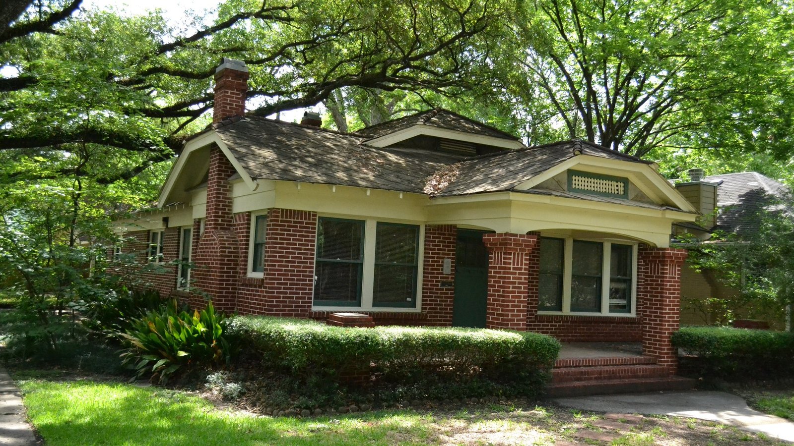 A red brick bungalow with a deep porch under large oak trees in Houston