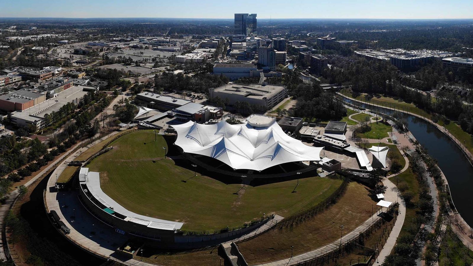 An aerial view of the Cynthia Woods Mitchell Pavilion canopy and lawn with The Woodlands Town Center buildings behind it