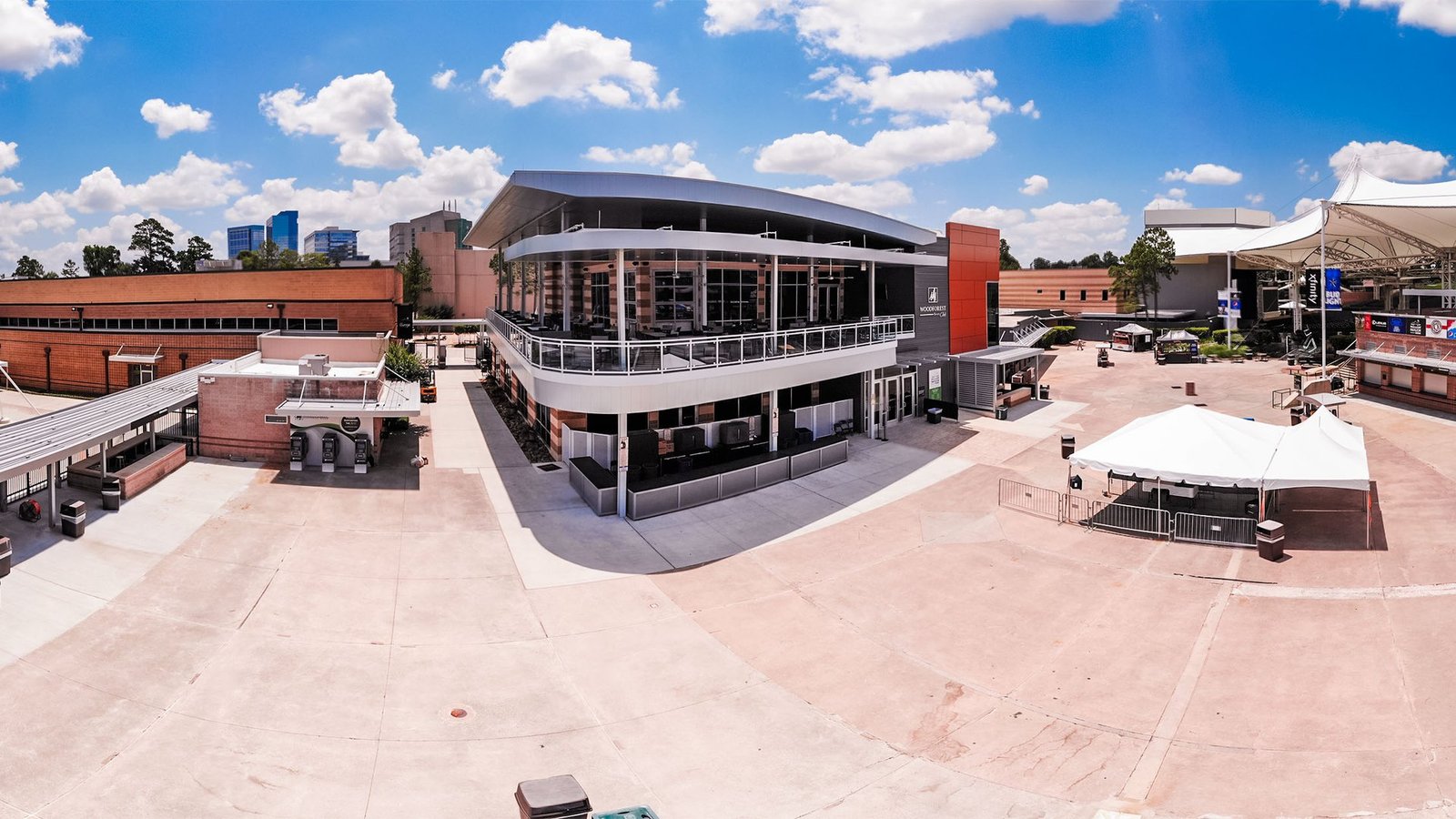 A wide sunny view of a paved plaza at the Cynthia Woods Mitchell Pavilion with a two-story glass building and white tents