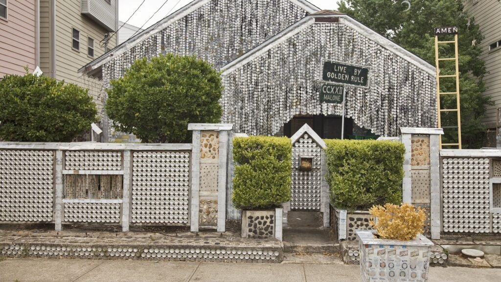 The front of the Beer Can House in Houston, its walls hung with flattened beer cans behind a can-covered fence and trimmed hedges