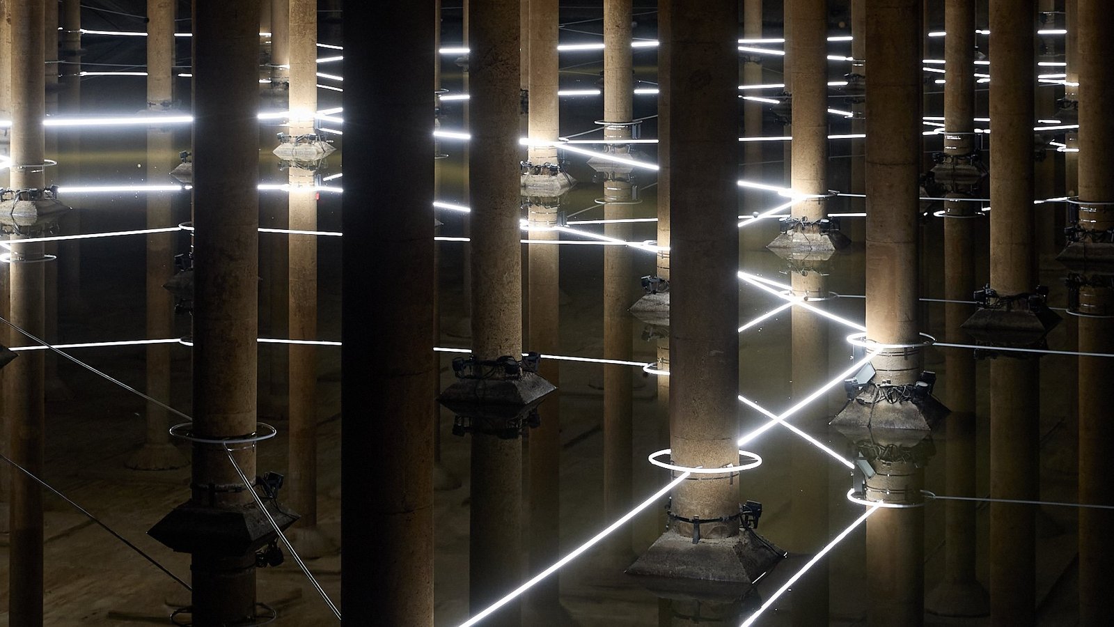 Rows of concrete columns inside the Buffalo Bayou Park Cistern, lit by white lines of light reflected in shallow water