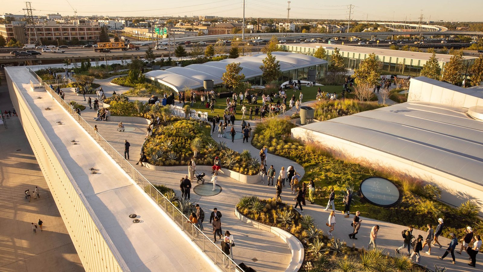 An aerial view of the Skylawn rooftop park at POST Houston, with curving paths, gardens and visitors at sunset